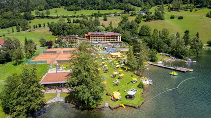 Luftaufnahme eines Ferienresorts am See mit Liegestühlen und Tennisplätzen