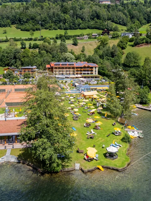 Luftaufnahme eines Ferienresorts am See mit Liegestühlen und Tennisplätzen