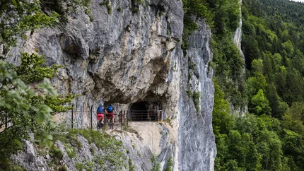 Two cyclists on narrow mountain path near rock cave and forest