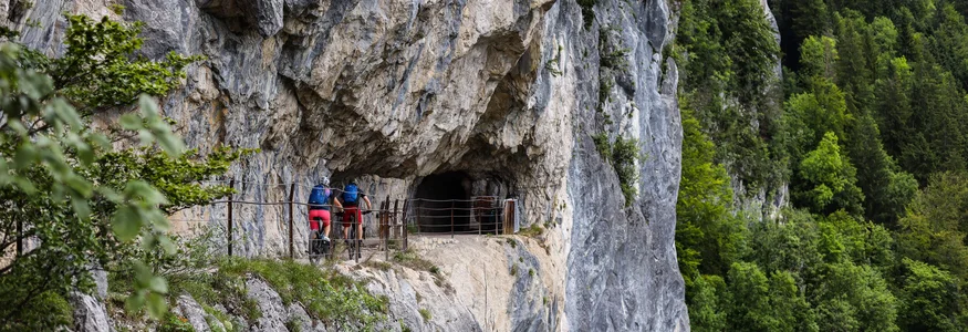 Twee fietsers op smal bergpad bij rotsachtige grot en bos