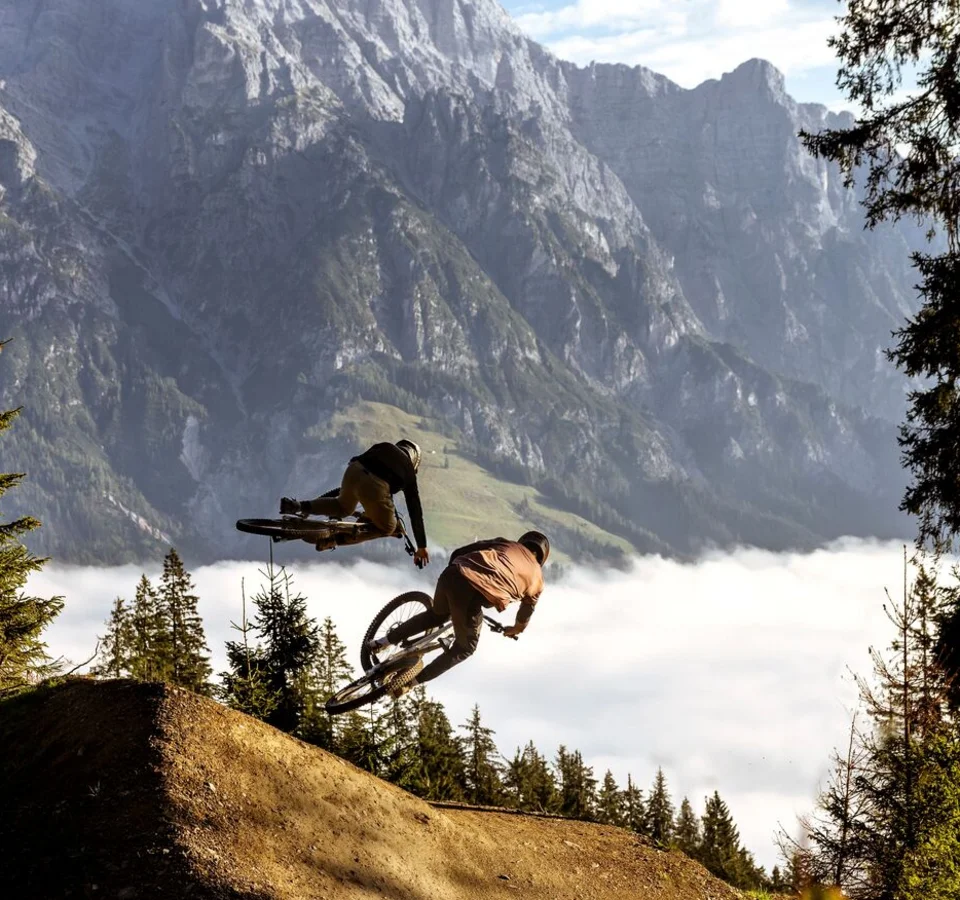 Bikepark Leogang © Klemens König Zwei Mountainbiker springen im Wald vor Bergkulisse über eine Rampe