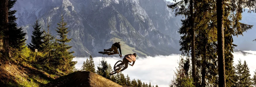 Bikepark Leogang © Klemens König Zwei Mountainbiker springen im Wald vor Bergkulisse über eine Rampe