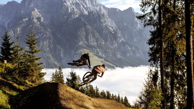 Zwei Mountainbiker springen im Wald vor Bergkulisse über eine Rampe