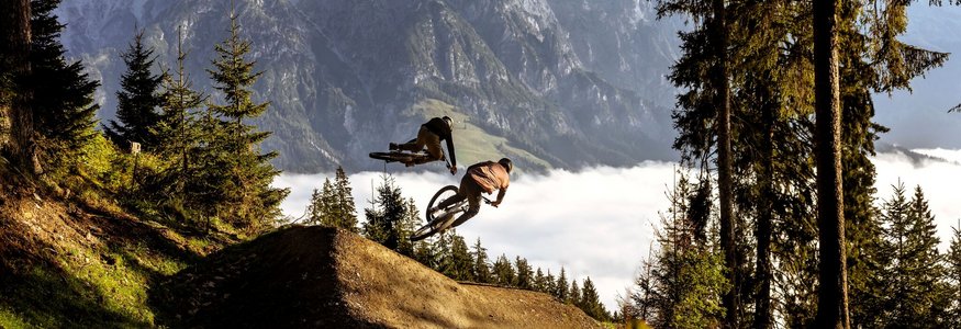 Zwei Mountainbiker springen im Wald vor Bergkulisse über eine Rampe