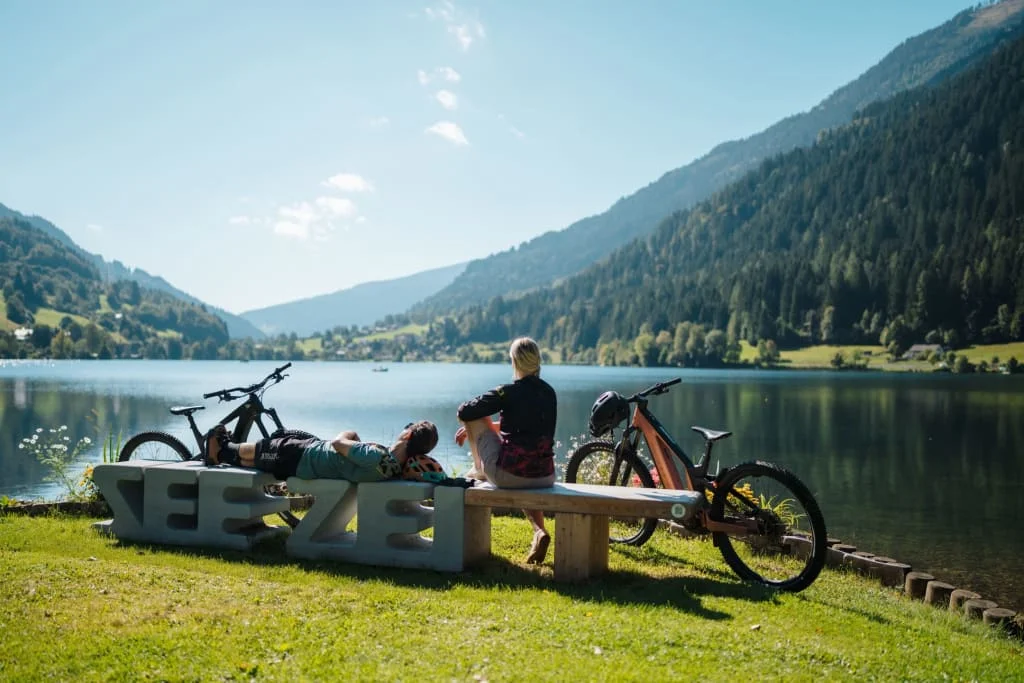 Zwei Personen mit Fahrrädern sitzen auf Bank am See vor Bergen