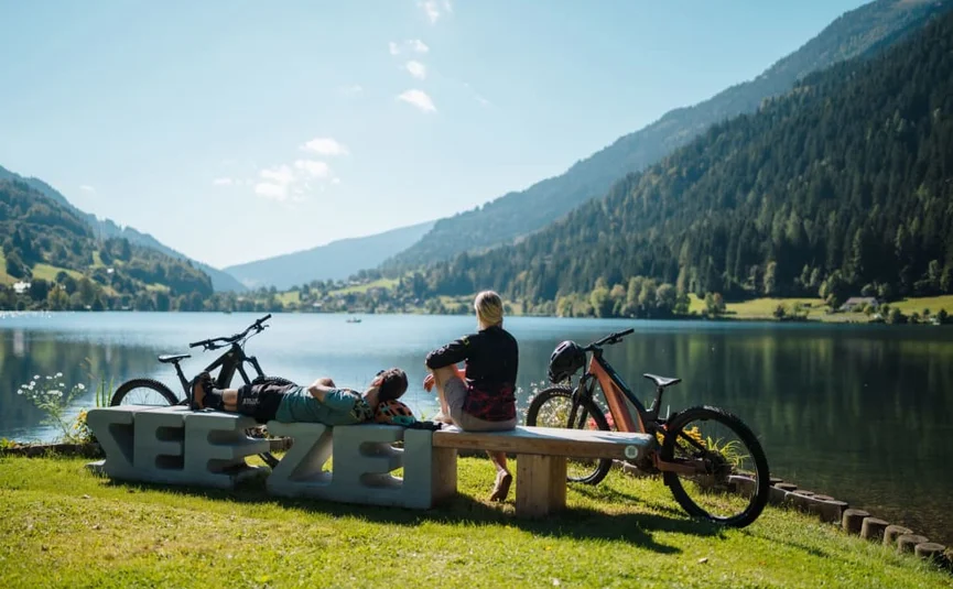 Zwei Personen mit Fahrrädern sitzen auf Bank am See vor Bergen