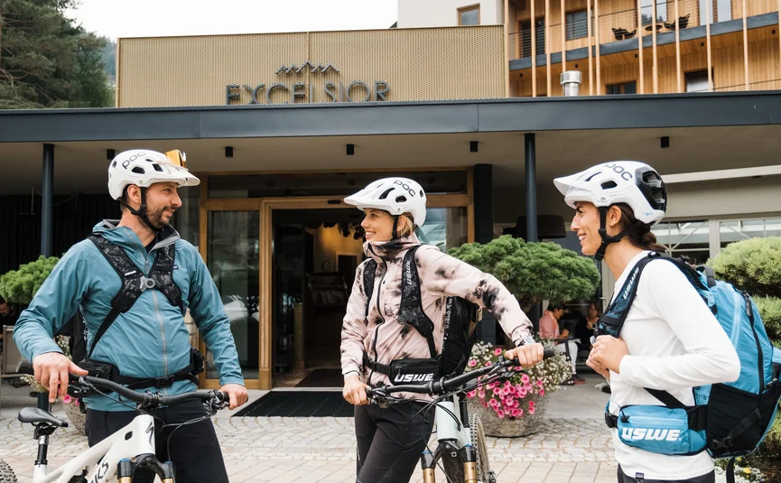 Three cyclists wearing helmets talking in front of Excelsior Hotel
