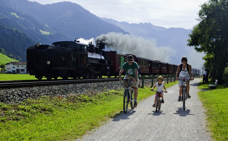 Family cycling beside a steam train in a mountainous landscape