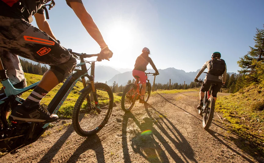 Drie mountainbikers fietsen op een zonnig bergpad met bergen op de achtergrond