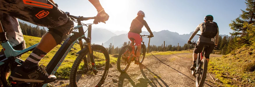 Brandnertal © Michael Marte - Bikepark Brandnertal Three mountain bikers riding on sunny mountain trail with mountains behind