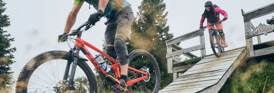 Zwei Mountainbiker fahren eine Rampe auf einem Waldweg hinunter