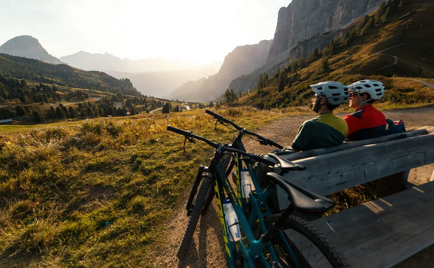 Two cyclists resting on a bench overlooking valley and mountains at sunset