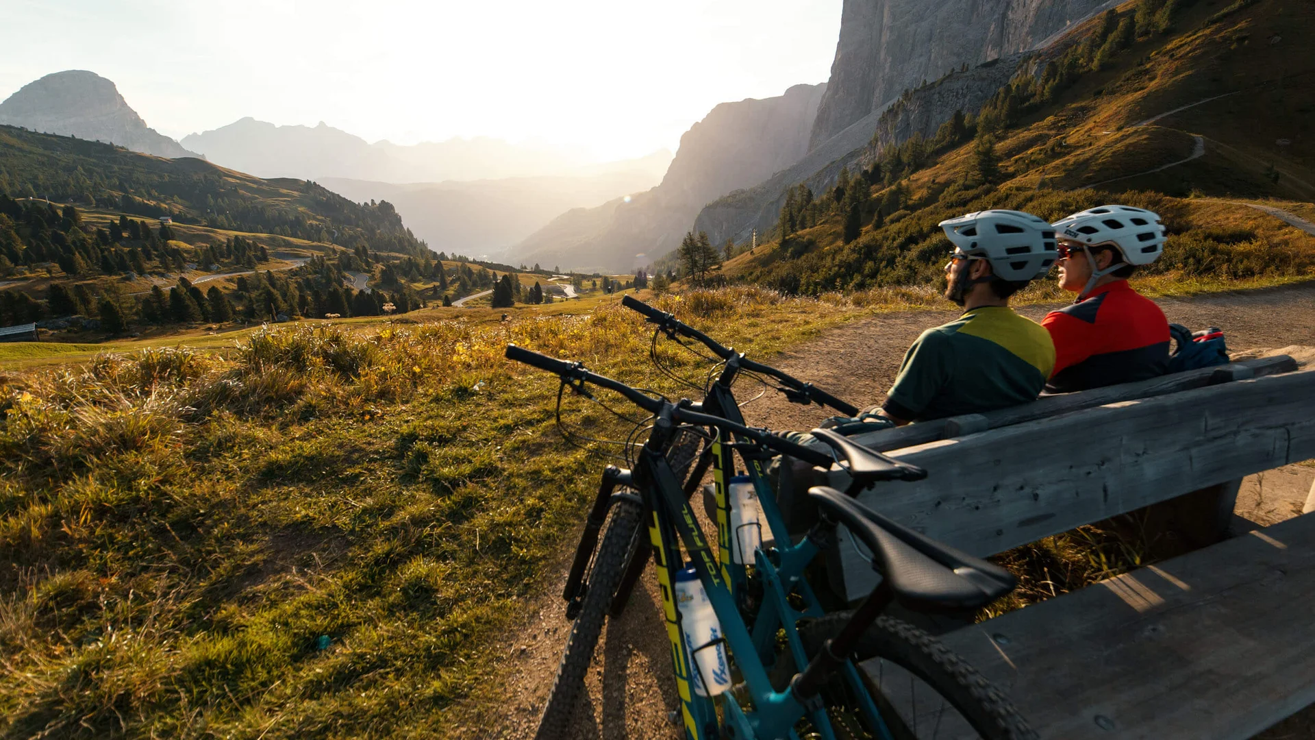 Zwei Radfahrer sitzen auf einer Bank und blicken auf das Tal und die Berge bei Sonnenuntergang