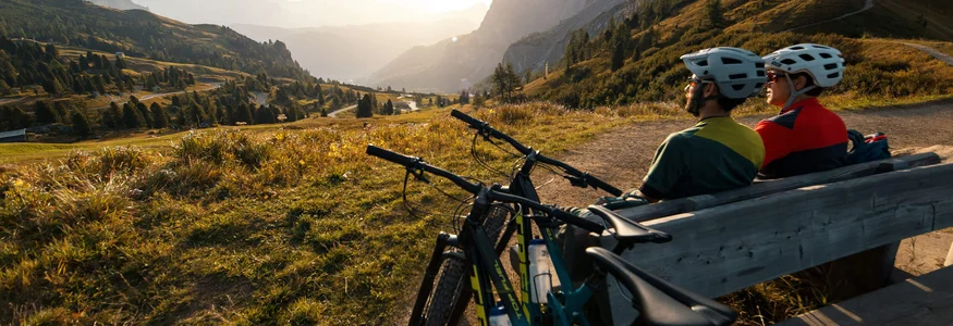 Two cyclists resting on a bench overlooking valley and mountains at sunset