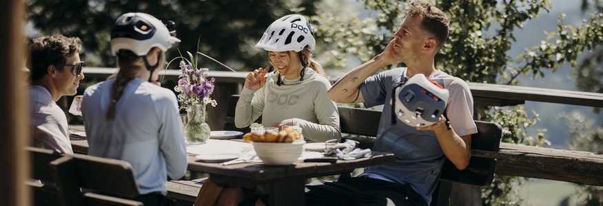 Gruppe von Radfahrern mit Helmen sitzt draußen an einem Holztisch und unterhält sich