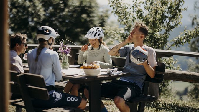 Gruppe von Radfahrern mit Helmen sitzt draußen an einem Holztisch und unterhält sich