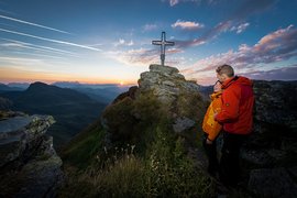 SUMMER IN THE MOUNTAINS Couple stands on mountain summit at sunset near summit cross