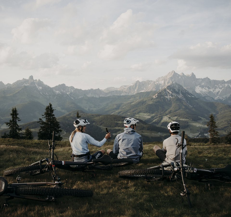 MTB in der DNA: Bikesommer im DIPS&DROPS © Matthias Warter Drei Radfahrer sitzen auf einer Wiese mit Mountainbikes vor Berglandschaft