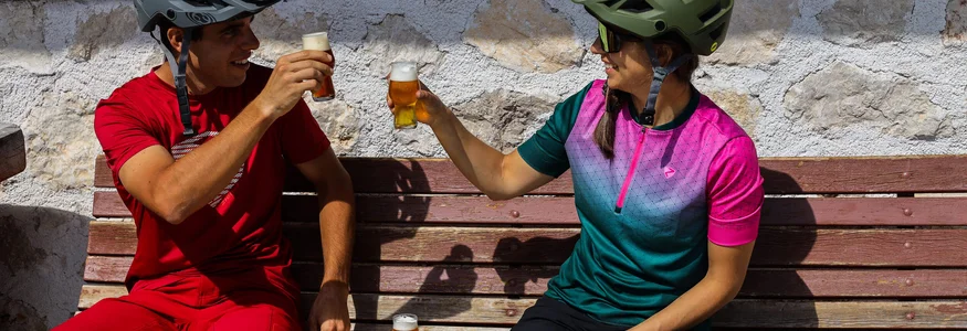 Zwei Radfahrer stoßen mit Biergläsern auf einer Holzbank an