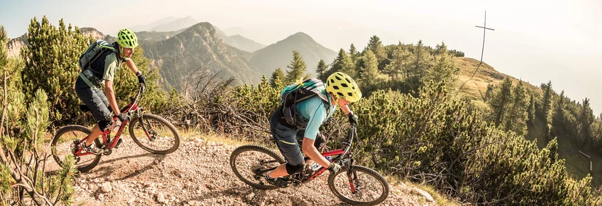 Zwei Mountainbiker fahren auf einem Bergpfad mit Bergen im Hintergrund.