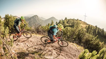 Two mountain bikers riding on a mountain trail with mountains in the background.
