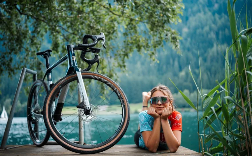 Woman lying on a dock with a bike by a lake and mountains behind