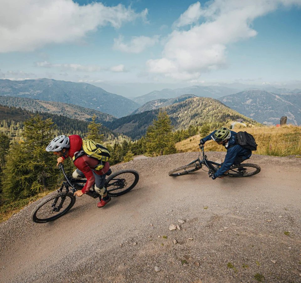 Two mountain bikers riding down a steep trail with mountains in the background