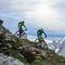 Zwei Mountainbiker fahren an einem steilen Berg mit schneebedeckten Gipfeln im Hintergrund