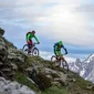 Twee mountainbikers dalen een steile heuvel af met besneeuwde bergtoppen op de achtergrond