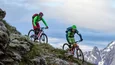Zwei Mountainbiker fahren an einem steilen Berg mit schneebedeckten Gipfeln im Hintergrund