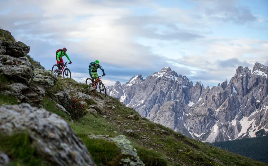 Twee mountainbikers dalen een steile heuvel af met besneeuwde bergtoppen op de achtergrond