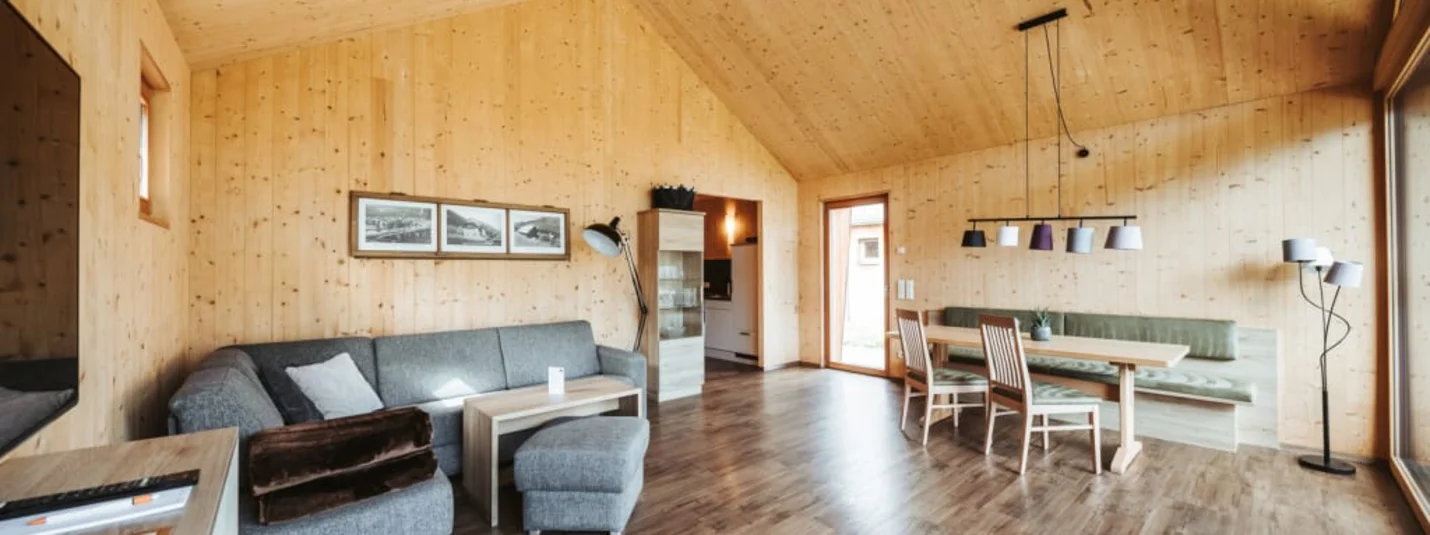 Gemütliches Wohnzimmer mit Holzwänden, grauem Sofa und Essbereich mit Tisch und Stühlen