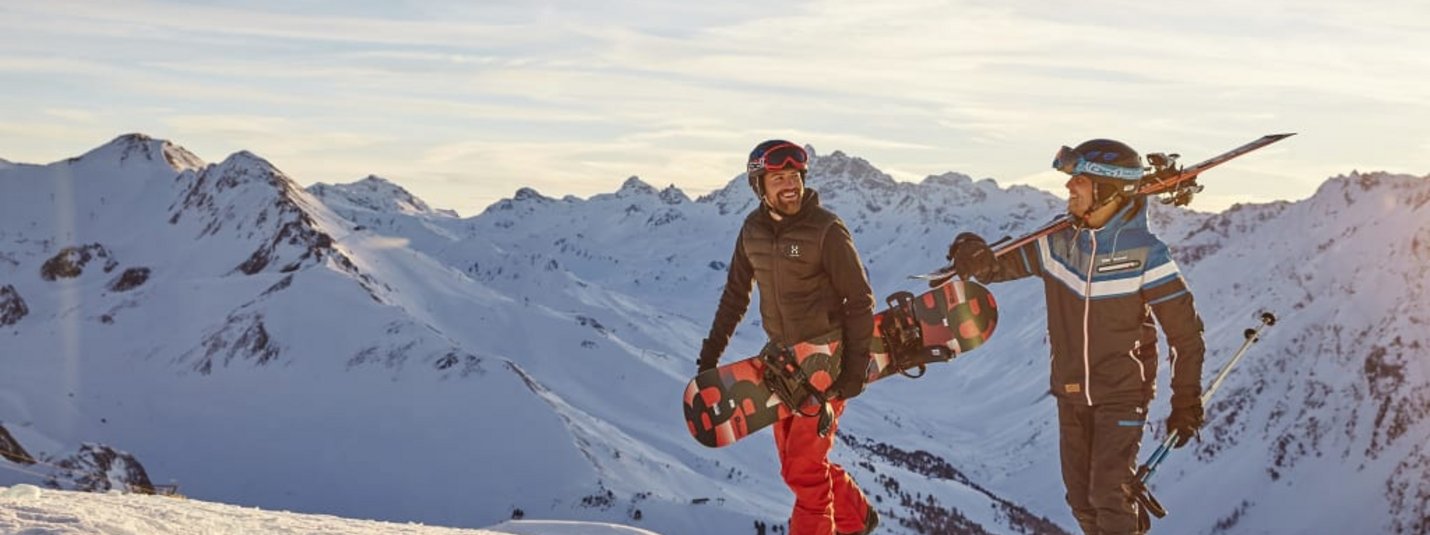 Zwei Skifahrer und Snowboarder wandern im Schnee mit Bergpanorama bei Sonnenuntergang