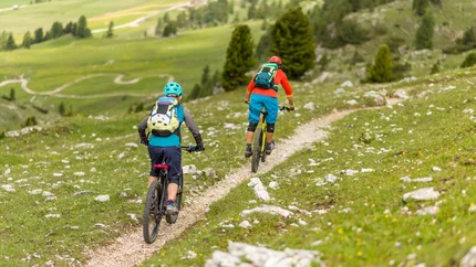 Two mountain bikers riding on a narrow trail through green mountainous terrain