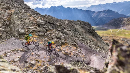 Zwei Mountainbiker fahren auf einem bergigen Pfad mit Alpen im Hintergrund