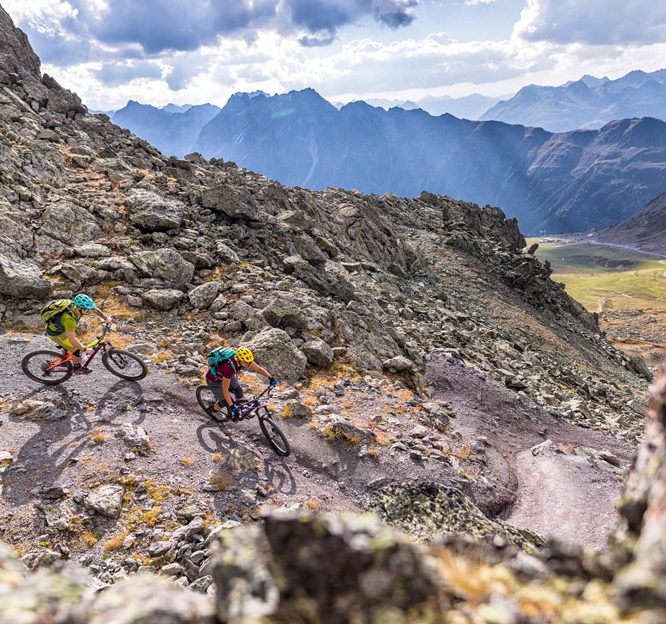 Two mountain bikers riding on a rocky trail in the mountains with alpine peaks