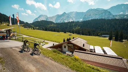 Zwei Mountainbiker fahren auf Bergweg mit Alpenhütte und Bergen im Hintergrund