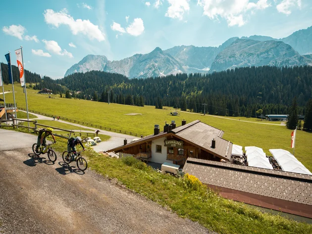 Zwei Mountainbiker fahren auf Bergweg mit Alpenhütte und Bergen im Hintergrund