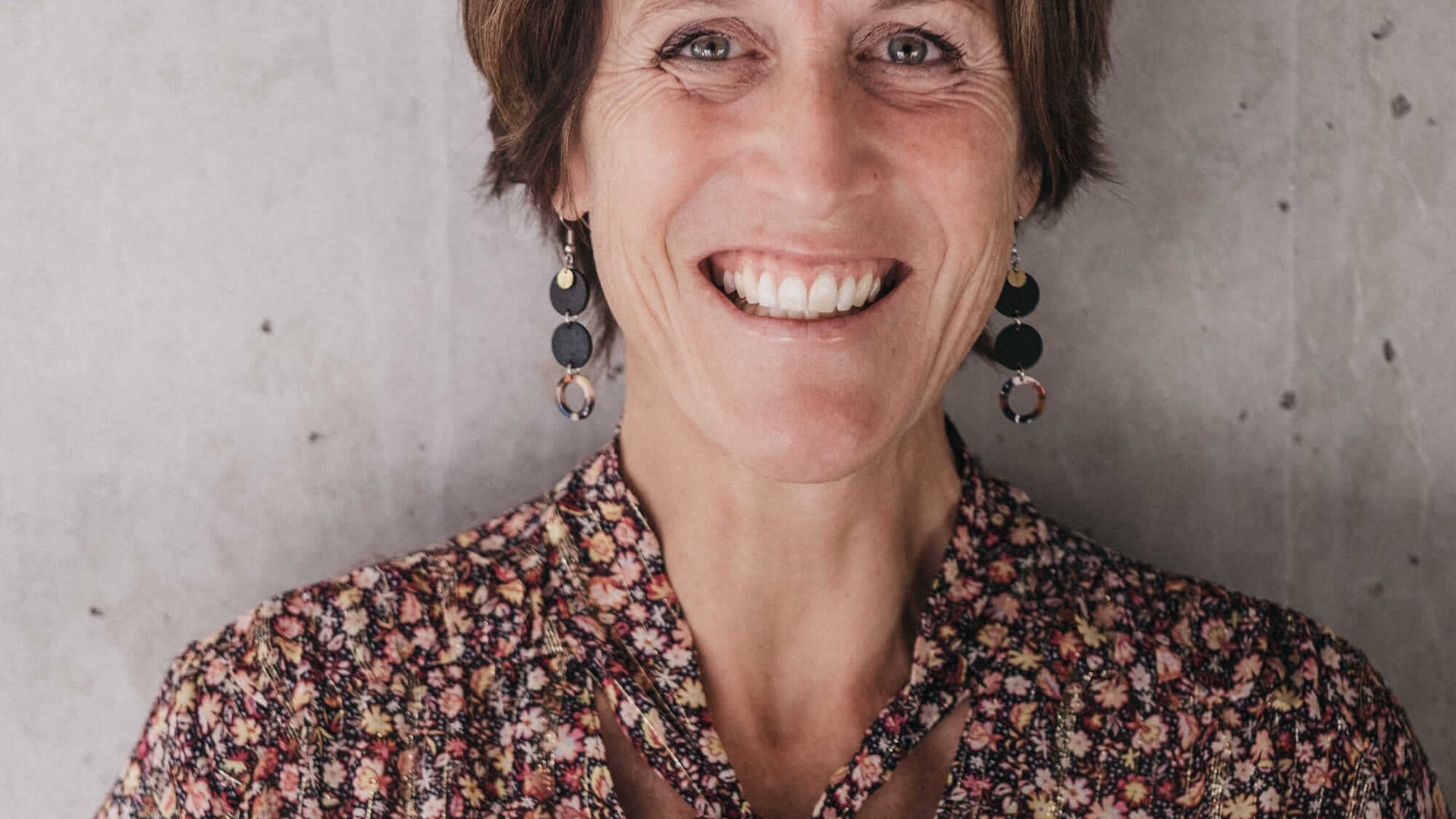 Hotel Rupertus: Ein Retreat für Yogis und Biker © Heldentheater Lächelnde Frau vor einer grauen Wand mit gemustertem Kleid und Ohrringen