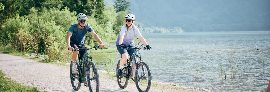 Two cyclists riding bikes on a lakeside path with trees in the background