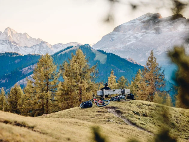 Mountainbike-Urlaub in Alta Badia © Tobias Köhler Person sitzt auf Bank in den Bergen mit zwei Mountainbikes und Wald im Hintergrund