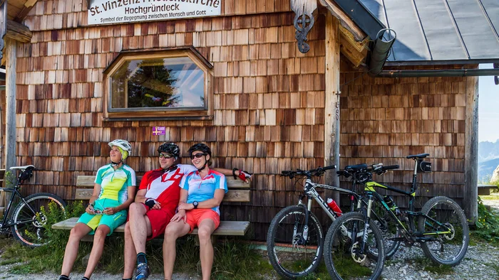 Bike-Region Flachau – von Radabenteuer bis Signature Ride © Salzburger Sportwelt - Coen Weesjes Drei Radfahrer sitzen auf einer Bank vor der St. Vinzenz Friedenskirche