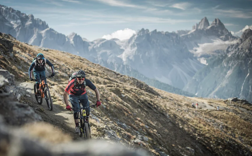Zwei Mountainbiker fahren auf einem Bergpfad mit Alpen im Hintergrund