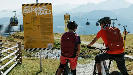 Zwei Mountainbiker lesen ein Schild an den Isskogel Trails mit Seilbahn im Hintergrund