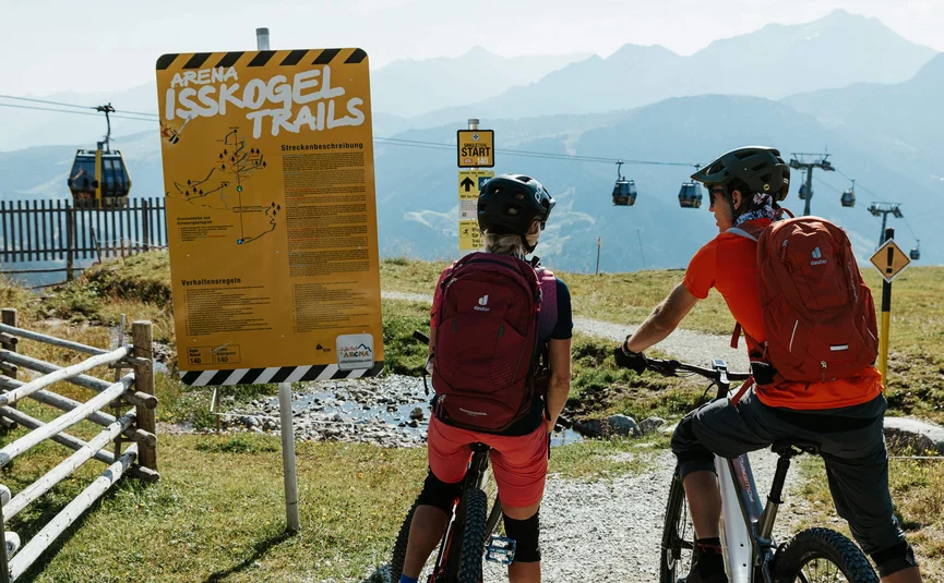 Zwei Mountainbiker lesen ein Schild an den Isskogel Trails mit Seilbahn im Hintergrund