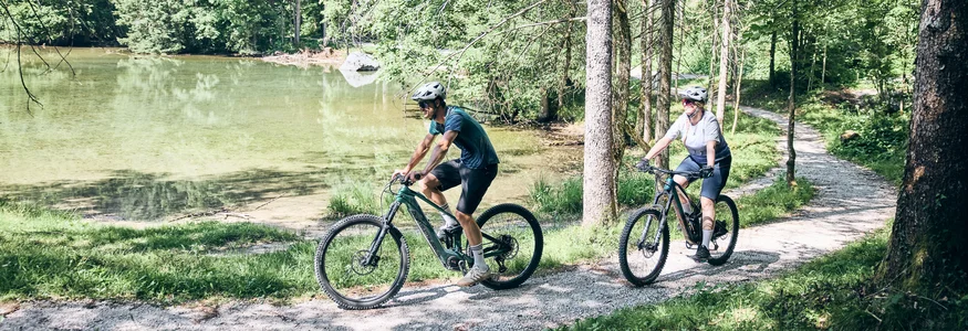 Two cyclists riding on a forest trail beside a lake on a sunny day