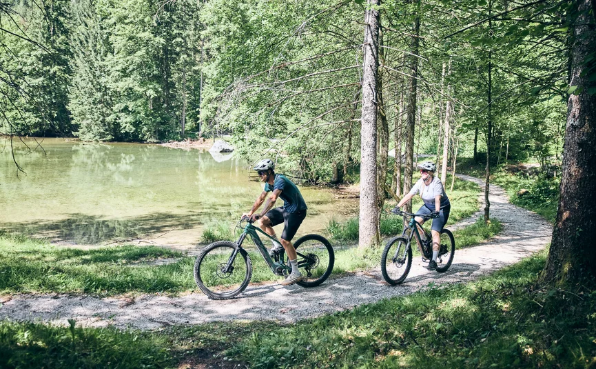 Two cyclists riding on a forest trail beside a lake on a sunny day