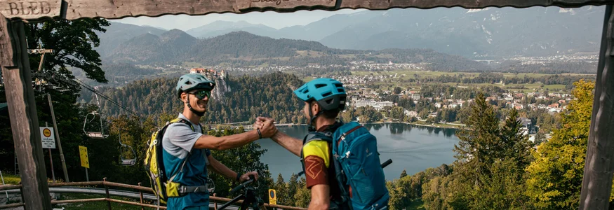 Dein Julische Alpen-Guide © Felix Saller Zwei Mountainbiker grüßen sich vor Alpenlandschaft mit See und Stadt