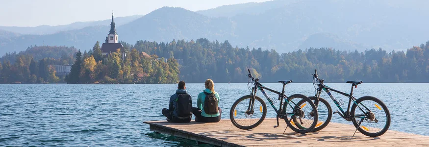 Zwei Personen sitzen mit Fahrrädern auf einem Steg am See mit Kirche und Bergen im Hintergrund