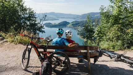 Twee fietsers zitten op een bankje met uitzicht op een meer en bergen.